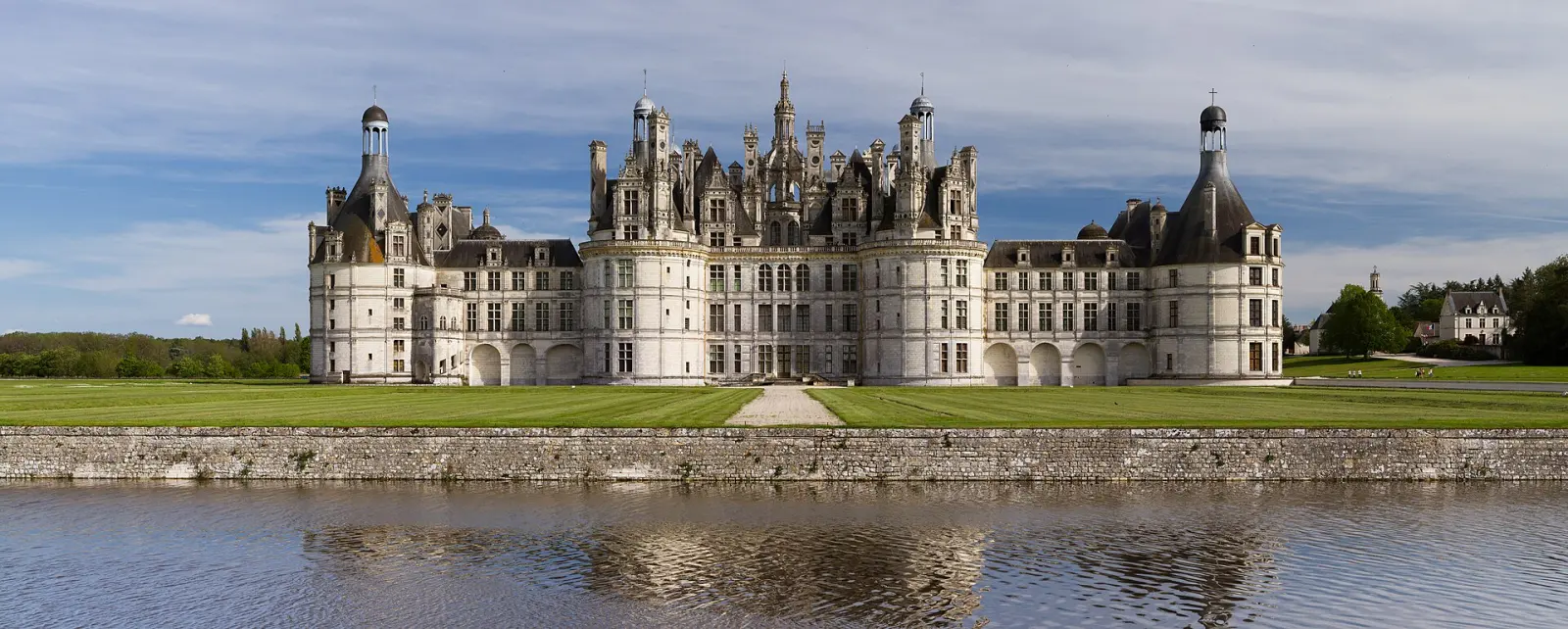 Le château de Chambord vu de la façade nord-ouest : un pavillon central à lanternons encadré de deux grandes tours rondes, devant une pelouse et un fossé en eau.